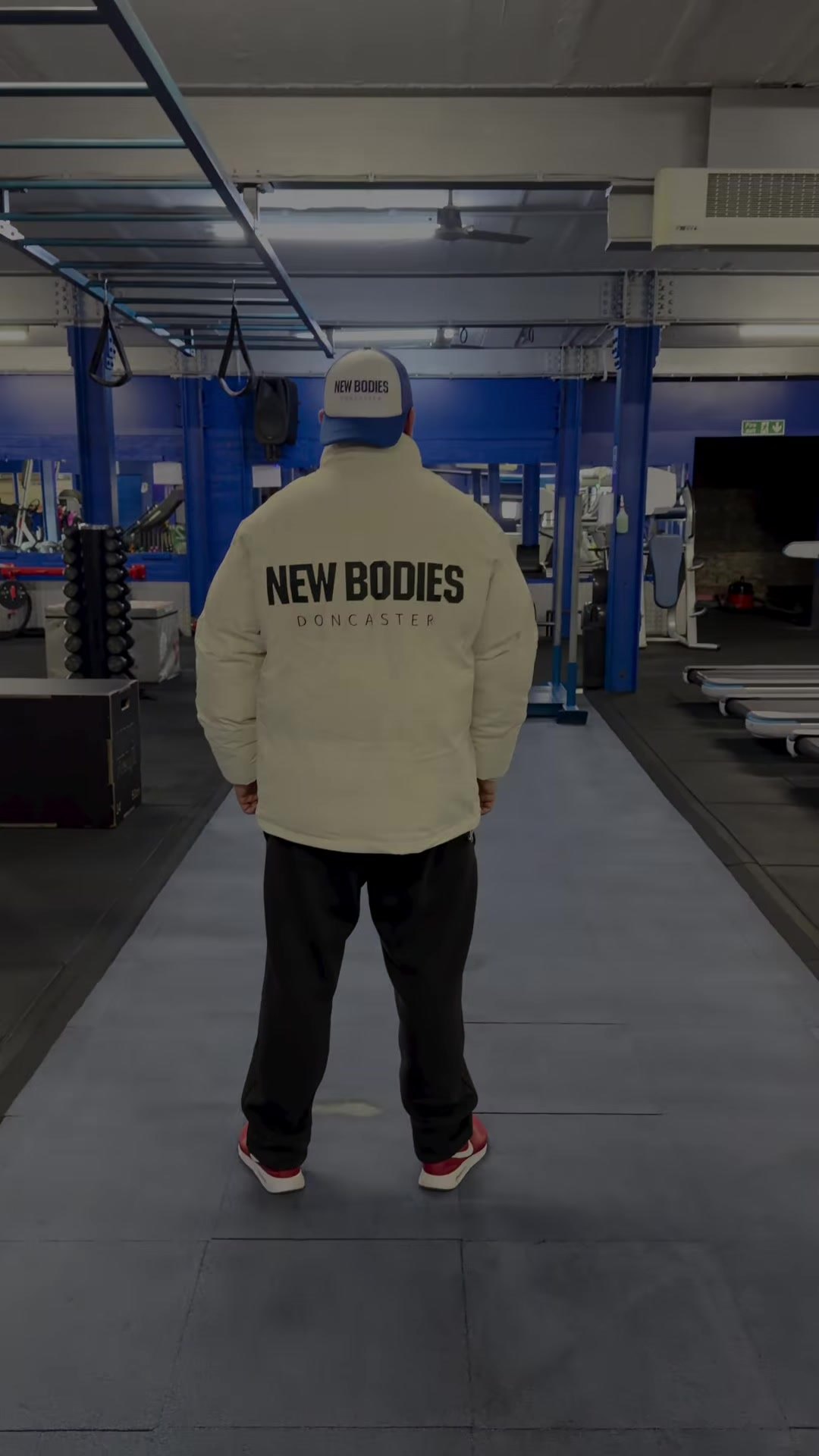 The New Bodies gym floor with blue pillars and a long black runway, a member in a cream New Bodies jacket seen from behind
